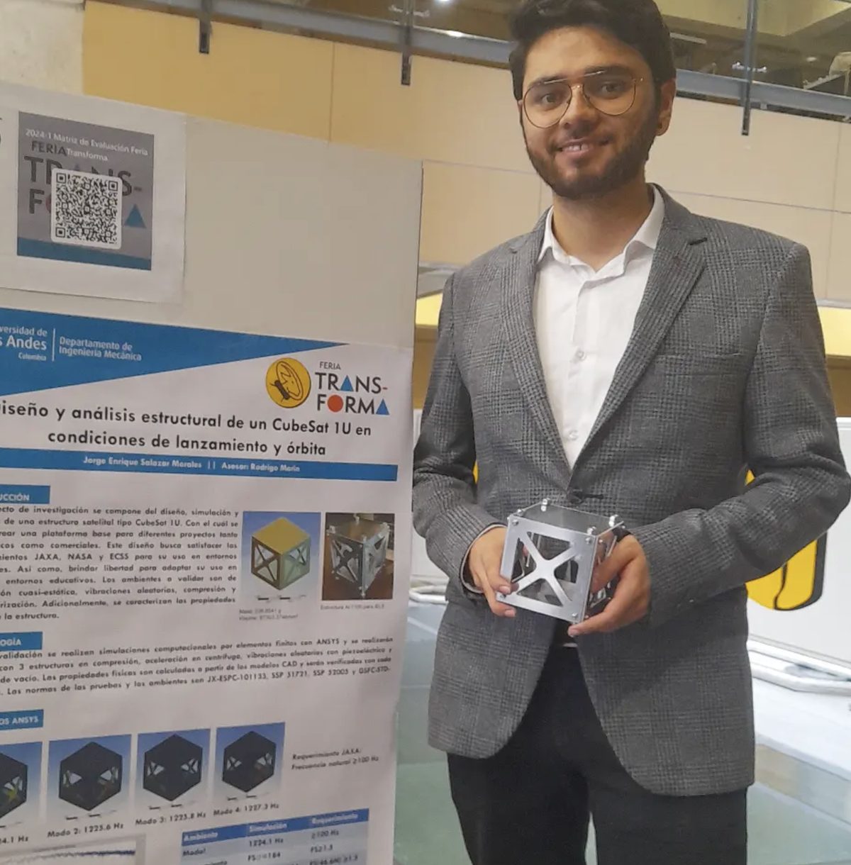 Estudiante sosteniendo la estructura de un CubeSat 1U junto al póster de su proyecto en la Feria Transforma